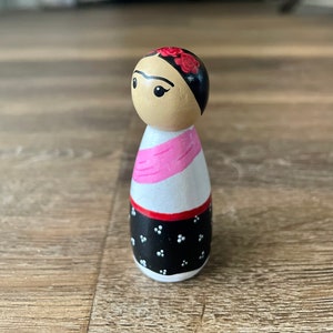 May include: A wooden doll painted to resemble Frida Kahlo. The doll has black hair, a pink scarf, and a black and white patterned dress. The doll is wearing a red flower crown.