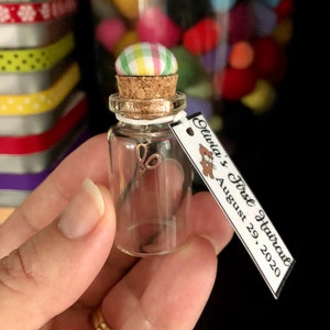 May include: A small glass bottle with a cork top and a fabric button on top. The bottle contains a pair of tiny scissors. A tag is attached to the bottle that reads "Olivia's First Haircut August 29, 2020".