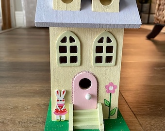 May include: A yellow wooden birdhouse with a purple roof, two small windows, and a pink door. The birdhouse has a green lawn base with a white bunny wearing a red dress and a pink flower.