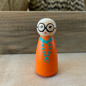 May include: A hand-painted wooden doll with white hair, black glasses, and a bright orange dress with a turquoise necklace.
