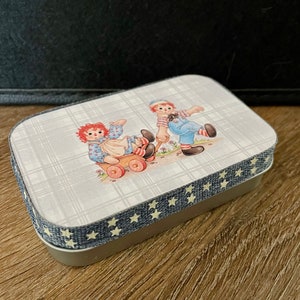 May include: A small, rectangular tin box with a blue and white plaid pattern and a white star trim. The lid features a vintage illustration of two rag dolls playing with a toy wagon.
