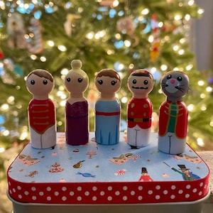 May include: A set of five wooden peg dolls in a blue and white tin with a red polka dot trim. The dolls are painted to look like characters from the Nutcracker ballet, including a ballerina, a nutcracker soldier, a mouse king, and a prince.