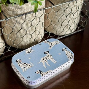 May include: A rectangular metal tin with a light blue lid featuring illustrations of dalmatian dogs and paw prints. The tin has a white dotted border and a silver base. The background includes a wire basket and potted plants.