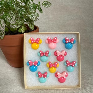 Op de afbeelding: Een verzameling kleurrijke Minnie Mouse-vormige accessoires, elk met een strik. De set bevat gele, roze en blauwe stukken met witte stippen op de strikken. Ze worden gepresenteerd in een kleine, vierkante doos.