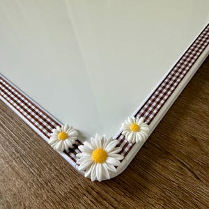 May include: A white dry-erase board featuring a brown and white gingham ribbon border along the bottom edge. Three white daisy embellishments with yellow centers are attached to the ribbon, with one larger flower centered between two smaller ones.