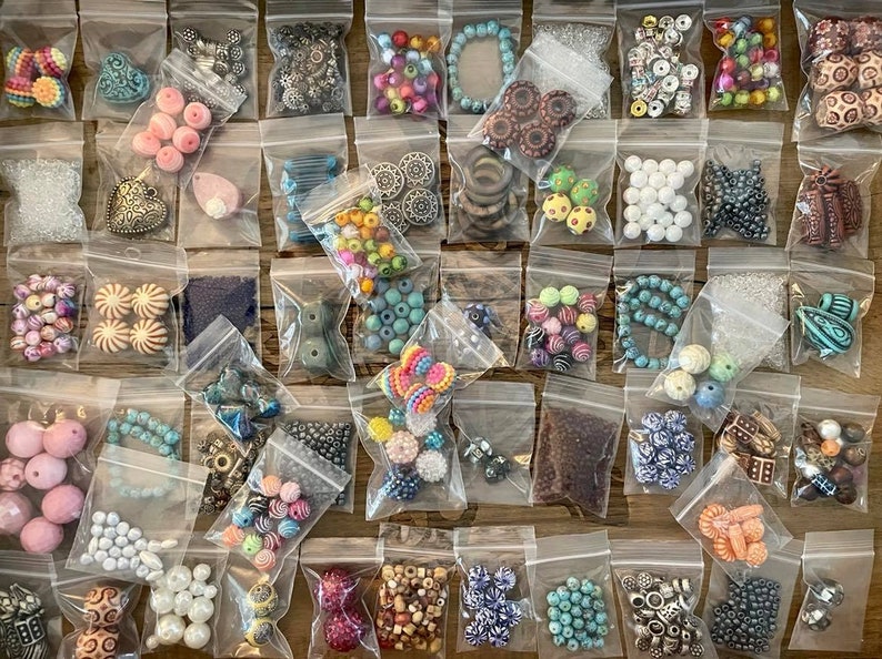 May include: Assortment of colorful beads and charms in small plastic bags. The bags are arranged in rows on a white surface. The beads are made of various materials, including glass, metal, and plastic. The charms are made of metal and have a variety of shapes and designs.