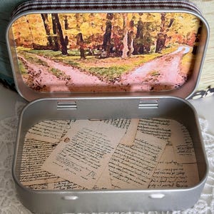 May include: A silver metal tin with a brown and white checkered fabric trim. The lid features a watercolor-style image of a path through a forest with fall foliage. The inside of the tin is lined with a patterned paper featuring handwritten text.