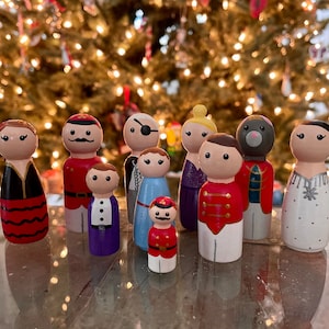 May include: A set of 10 hand-painted wooden peg dolls, each with unique features and clothing. The dolls are dressed in various costumes, including a red and black dress, a blue and white sailor suit, and a white and gold dress. The dolls are perfect for imaginative play and storytelling.