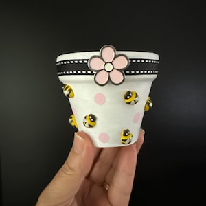 May include: A small white flower pot decorated with pink polka dots and cartoon bees. A black ribbon with white stitching encircles the top, and a pink flower is attached to the front.