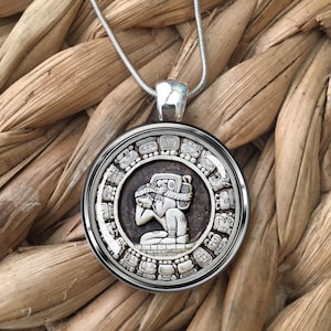 May include: A silver chain necklace with a round pendant featuring a detailed engraving of a Mayan calendar with a figure in the center.