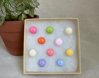 May include: A small, open box filled with colorful, round beads. The beads are in shades of pink, green, orange, white, light pink, red, yellow, purple, and blue. A potted plant with green and white speckled leaves is in the background.