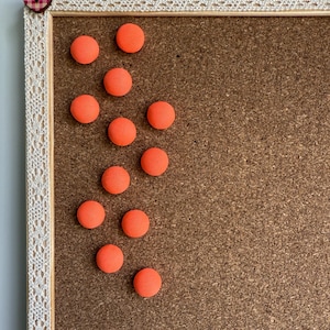 May include: A corkboard with a wooden frame, adorned with a white lace trim and a heart-shaped button. Bright orange circular magnets are scattered across the board. The corkboard is a practical and decorative item.