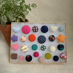 May include: A collection of colorful fabric-covered buttons in various shapes and patterns, including polka dots, gingham, and floral designs. The buttons are arranged in a rectangular box, with a potted plant in the background.