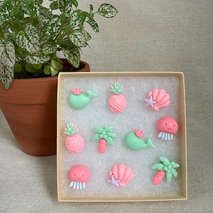 May include: A collection of miniature, pastel-colored ocean-themed figurines in a box. The set includes pink pineapples, green whales with pink crowns, pink jellyfish, seashells with purple starfish, and palm trees. A potted plant is in the background.