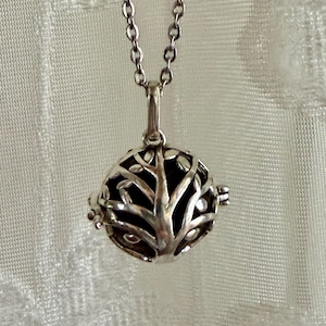 May include: A silver-toned necklace featuring a round pendant with a tree-of-life design. The pendant has an openwork pattern, revealing a dark interior. The chain is delicate and hangs from the pendant's top.
