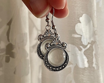 May include: Silver earrings with a white, round, opaque stone set in a decorative, ornate frame.