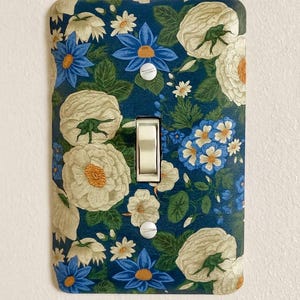 May include: Decorative light switch cover featuring a floral pattern. The cover has a dark blue background with white and blue flowers and green leaves. The switch is white.