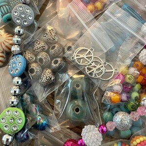 May include: Assortment of colorful beads and charms in small plastic bags. The beads are various shapes, sizes, and colors, including silver, turquoise, pink, green, and white. Some of the charms are peace signs.