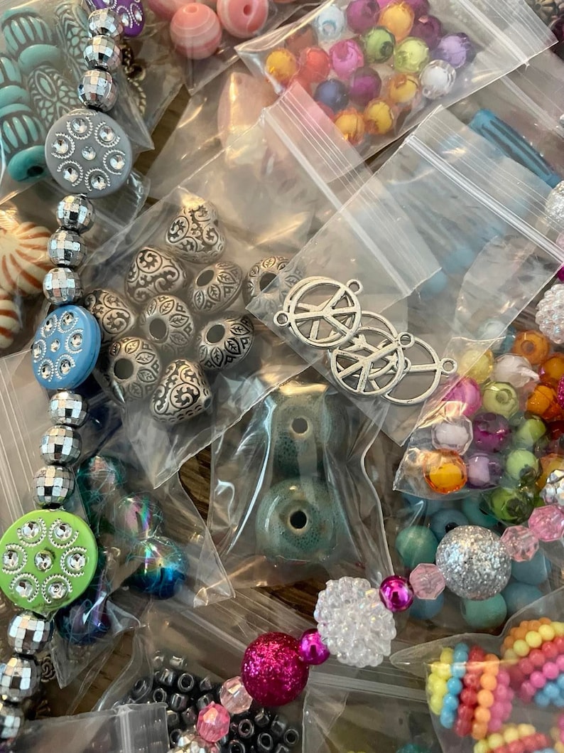 May include: Assortment of colorful beads and charms in small plastic bags. The beads are various shapes, sizes, and colors, including silver, turquoise, pink, green, and white. Some of the charms are peace signs.