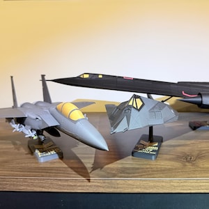 May include: Three model airplanes are displayed on a wooden shelf. A gray F-15 Eagle, a black SR-71 Blackbird, and a gray F-117 Nighthawk are visible. The F-117 Nighthawk has the text "ELITE SQUADRON F-117 NIGHTHAWK" on its base.