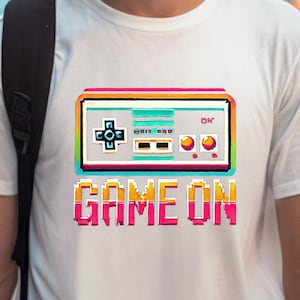 Könnte beinhalten: Weißes T-Shirt mit einem pixelartigen Bild eines Retro-Videospiel-Controllers und dem Text "GAME ON" in Regenbogenfarben.