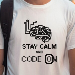 Puede incluir: Camiseta blanca con un gráfico de cerebro pixelado negro y el texto "Stay Calm and Code On".