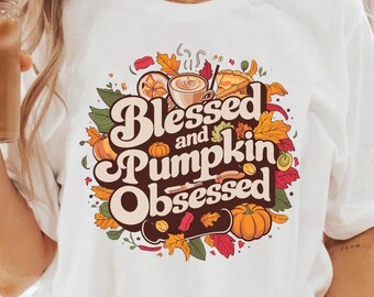 May include: White t-shirt with a graphic design of fall leaves, a pumpkin, and a coffee cup. The text on the shirt reads "Blessed and Pumpkin Obsessed".