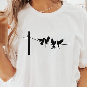 Könnte beinhalten: Weißes T-Shirt mit einem Motiv von Vögeln auf einer Wäscheleine. Das Shirt wird von einer Person getragen, die auf einer weißen Oberfläche sitzt. Die Person trägt weiße Socken mit schwarzen Streifen.