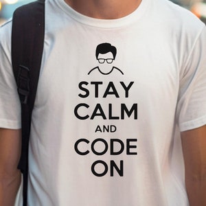 May include: White t-shirt with a black graphic that says "Stay Calm and Code On". The graphic also includes a simple black silhouette of a person wearing glasses.