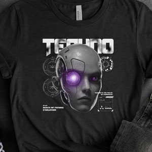 Puede incluir: Camiseta negra con un gráfico de una cara de robot con un ojo morado y la palabra "TECHNO" encima. La cara del robot está rodeada de engranajes y otros detalles técnicos. La camiseta está colocada sobre una superficie gris con un par de zapatillas Converse negras y un par de jeans de lavado oscuro.