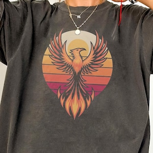May include: An oversized t-shirt in dark grey with a vintage-style graphic of a phoenix rising from flames against a sunset background.