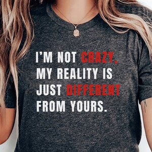 Może przedstawiać: Szary T-shirt z napisem "Nie jestem szalona, moja rzeczywistość jest po prostu inna niż twoja." w białych literach. Słowo "szalona" jest w czerwonych literach. Koszulkę nosi osoba z długimi blond włosami.