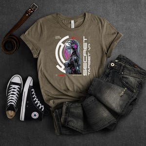 May include: A brown t-shirt with a graphic of a woman with pink hair and a futuristic design. The text "SECRET TARGET" is printed on the shirt.