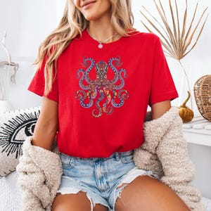 May include: A red t-shirt featuring a detailed, colorful octopus design. The octopus is depicted with intricate patterns in shades of blue, yellow, and orange. The shirt is a vibrant red, and the overall style is casual and artistic.