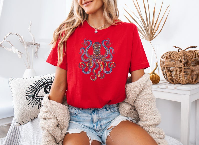 May include: A red t-shirt featuring a detailed, colorful octopus design. The octopus is depicted with intricate patterns in shades of blue, yellow, and orange. The shirt is a vibrant red, and the overall style is casual and artistic.