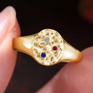 May include: A gold ring with an oval face, featuring three small gemstones: a light blue, a dark blue, and a red. The ring has a textured surface on the face and a smooth, polished band. The ring is held in a hand.