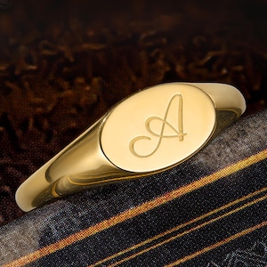 Peut inclure: Une bague chevalière en or poli présentant une face ovale gravée de la lettre A en écriture cursive. La bague possède une surface métallique lisse et réfléchissante ainsi qu'un anneau effilé, illustrant un bijou classique avec une initiale.