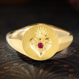 May include: A gold signet ring featuring a brushed finish on the circular face. The center of the face displays an engraved Sacred Heart motif with a small, round, red gemstone set directly in the middle of the heart design.