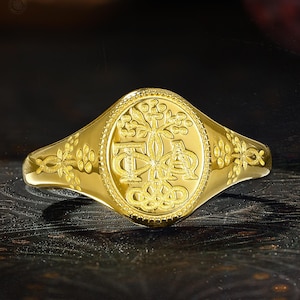 May include: A gold signet ring featuring an oval face with an engraved tree motif and the letters Phi and Alpha. The band displays intricate floral detailing on both sides, with a polished finish and a beaded border surrounding the central design.