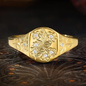 May include: A gold signet ring featuring an oval face engraved with a floral pattern and inlaid with six small clear stones. The band and the face of the ring are decorated with additional engraved leaf and vine motifs on both sides.