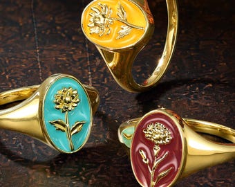 Könnte beinhalten: Drei goldene Siegelringe mit ovalen Zentren und Emaille-Einlagen. Ein Ring hat eine gelbe Einlage mit einer goldenen Blume, einer hat eine türkisfarbene Einlage mit einer goldenen Blume und einer hat eine dunkelrote Einlage mit einem Blumenmotiv.