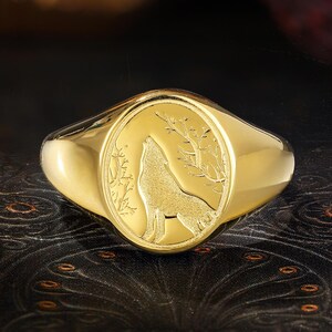 May include: A gold signet ring with an oval face. The face features a detailed engraving of a howling wolf and foliage. The ring has a polished finish and a thick band.