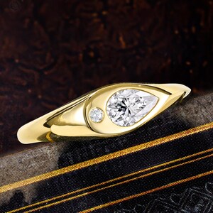 May include: A gold ring featuring a pear-shaped clear gemstone and a smaller round clear gemstone set side-by-side in a smooth, polished metal band. The design incorporates two distinct stone shapes within a single continuous gold setting.