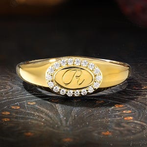 May include: A gold ring with an oval centre featuring the letter "R" and surrounded by small, clear gemstones. The ring has a polished, smooth band and a bright, reflective surface. The ring is likely made of gold and is a piece of jewellery.
