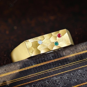 May include: A gold ring with a textured surface and three gemstones: a light blue, a red, and a green. The ring has a rectangular top and a smooth band. The ring is displayed on a dark, patterned surface.