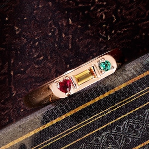 May include: A gold ring with a heart-shaped ruby, a rectangular yellow gemstone, and a triangular green emerald. The ring is set against a dark, patterned background.