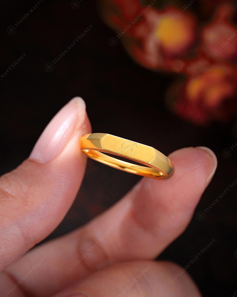 May include: A gold-colored ring with a faceted design. The ring has a single initial engraved on the top surface. The ring is held between two fingers against a dark background. The ring is a piece of jewelry.