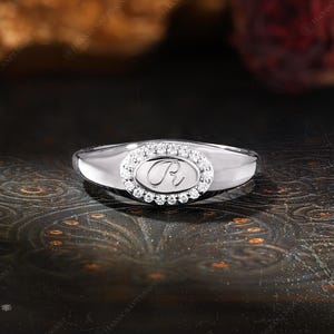 May include: A silver ring with an oval-shaped center stone. The center stone is surrounded by small, clear stones and features the letter "R". The ring has a polished finish and a classic design.