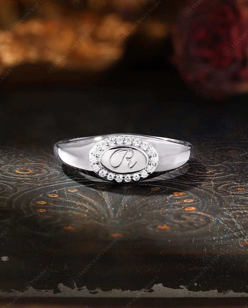 May include: A silver ring with an oval-shaped center stone. The center stone is surrounded by small, clear stones and features the letter "R". The ring has a polished finish and a classic design.