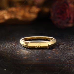 May include: A gold signet ring with an engraved "R" on a dark background. The ring has a geometric design with a flat top and beveled edges. The ring is likely made of gold and is a piece of jewelry.